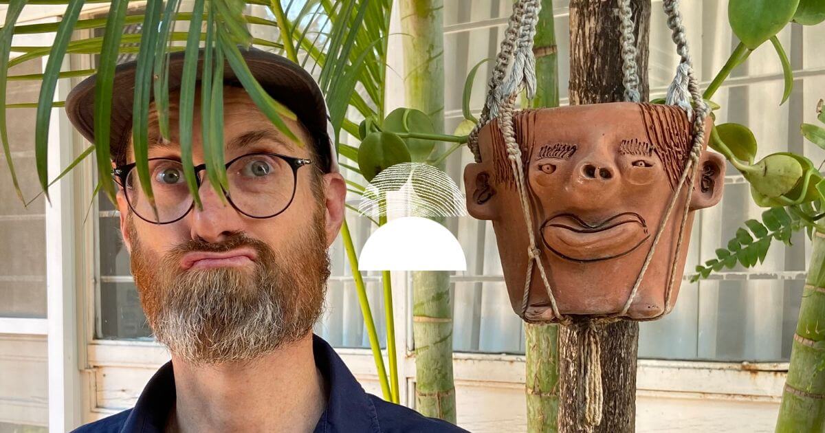A photo of our brand designer Rob making faces next to a plant pot with a funny face with green leaves and two semi-circles one solid white and the other one with wood grain marking